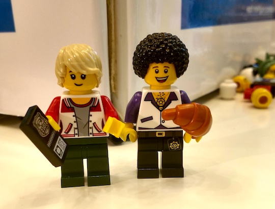 Two LEGO men, one blond in a red letter jacket holding a police badge, and one with dark curly hair in an open-collared disco shirt holding a croissant.